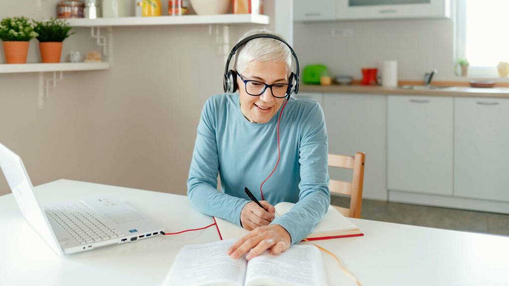 Aikuinen istuu tietokoneen ja kirjan äärellä keittiössä kuulokkeet päässä.